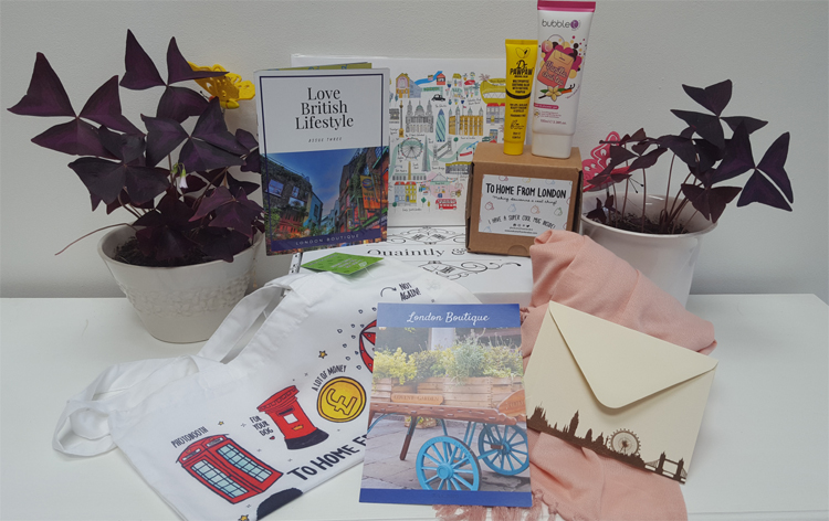 white gift box with the contents spread out in between two purple plants. the contents include a magazine, mug in a box, tote bag showing a london phone box and post box, a pink shawl and a leaflet picturing a cart with plants on top